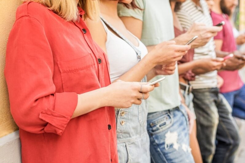 Teens are standing next to each other while looking at their phones.