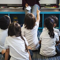 Children are sitting on the floor in a classroom looking at a teacher.