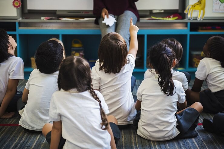 Children are sitting on the floor in a classroom looking at a teacher.