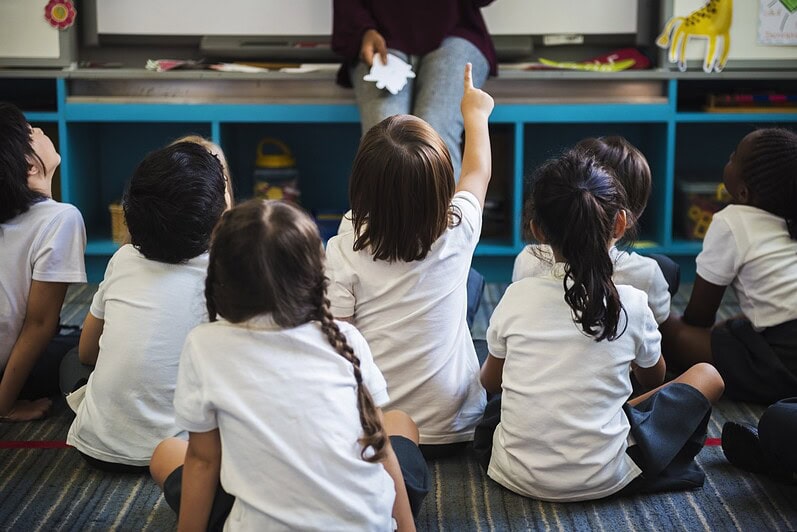 Children are sitting on the floor in a classroom looking at a teacher.