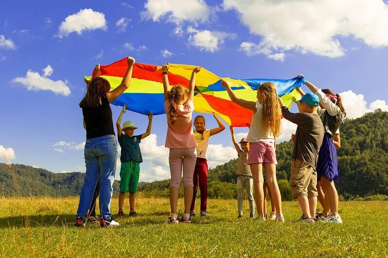 A group of children outdoors play under a multi-colored parachute.