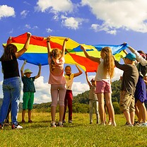 A group of children outdoors is playing under a multi-colored parachute.