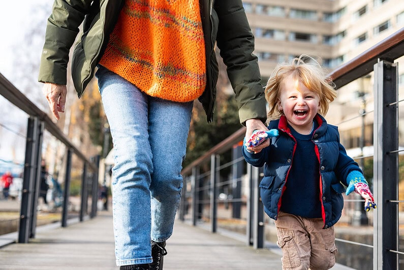 A happy toddler is walking outdoors while holding her parent's hand.