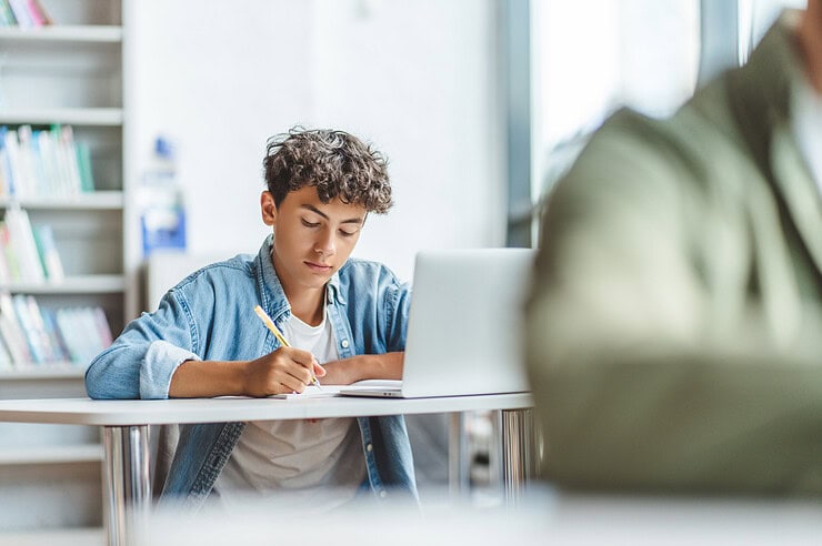 A teenager is completing a written exam, while his laptop is open on his desk.
