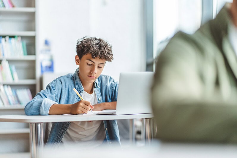 A teenager is completing a written exam, while his laptop is open on his desk.