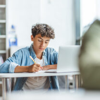 A teenager is completing a written exam, while his laptop is open on his desk.