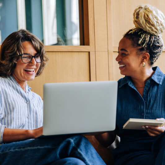 Two smiling teachers are having a friendly discussion while seated in front of an open laptop.