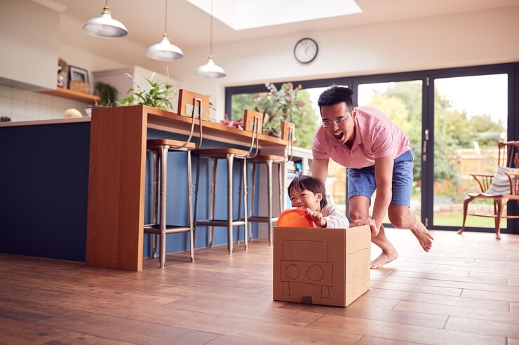 A father pushes his child in a box which they pretend is a car.