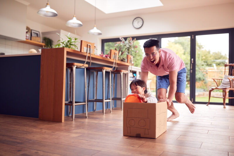 A father pushes his child in a box which they pretend is a car.
