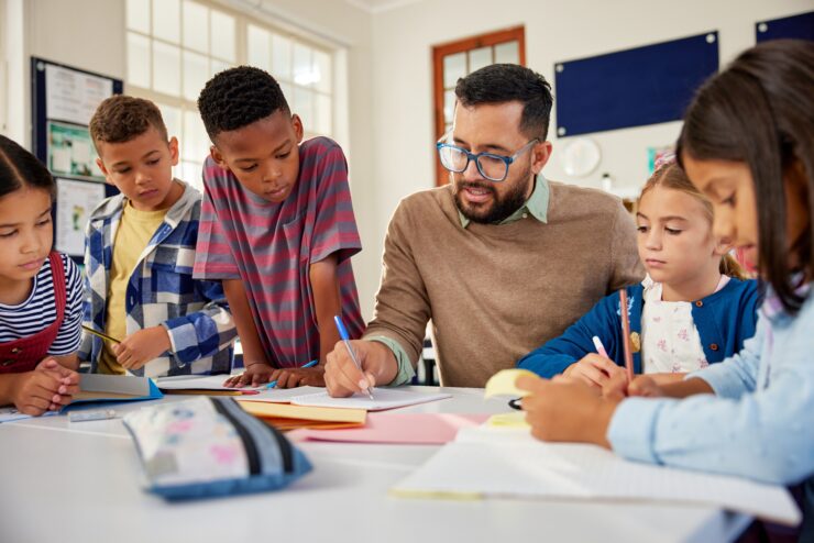 A teacher helps a group of young students with their schoolwork at a classroom table.