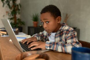 A boy types on a laptop while glancing sideways cheekily.