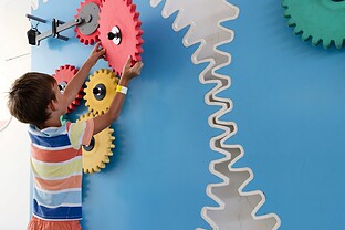A child is interacting with colorful gears on a mechanical wall.