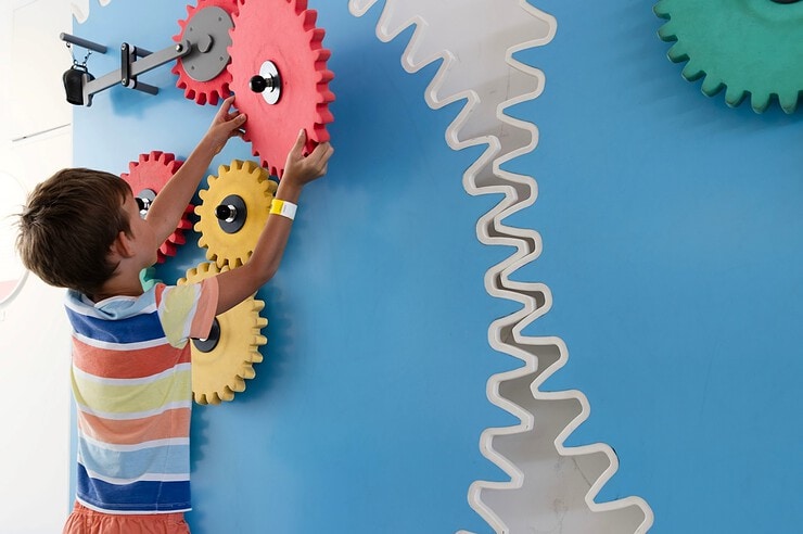A child is interacting with colorful gears on a mechanical wall.