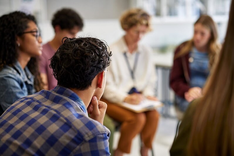 An adult and teenagers are sitting in a circle having a discussion.