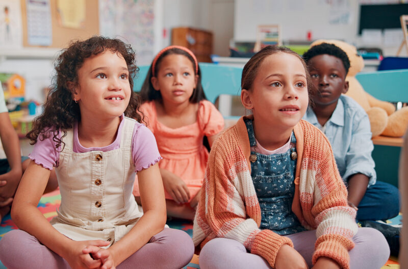 Children sitting on the classroom carpet look attentive.